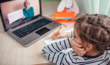 A girl writes on a notepad while watching a teacher on a laptop screen