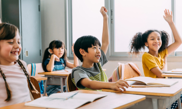 Four children sit smiling at desks in a classroom, with their hands raised in the air