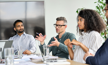 Three people sit at a conference table. The person in the middle is speaking