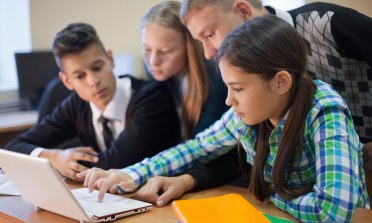 Three teenagers look at a laptop on a desk with their teacher