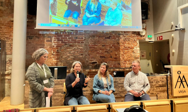 Four people sit in front of a presentation screen, one of them is speaking into a microphone