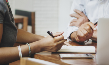 Two people's hands are holding pens as they take notes on a notepad