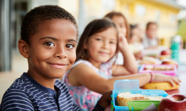 Children sit in a row, smiling at the camera, with their lunchboxes open on the table in front of them