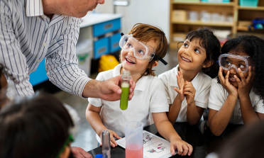 Children wearing safety goggles smile up at their teacher who is holding a test tube