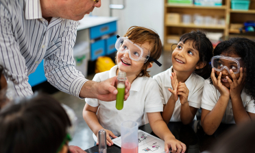 Children wearing safety goggles smile up at their teacher who is holding a test tube