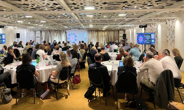 Participants at the bi-annual meeting sit around round tables and watch a presentation on a screen at the front of the room