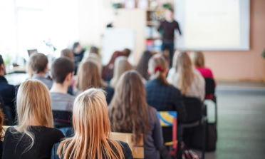 A group of people sit in rows, listening to someone speaking at the front of the room