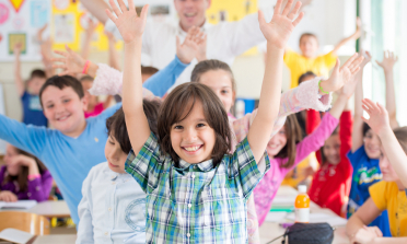 chidren in a classroon smiling and holding their hands high