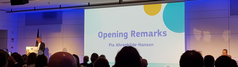 A screen which reads 'Opening remarks', with Pia Ahrenkilde Hansen standing at a lectern to the left