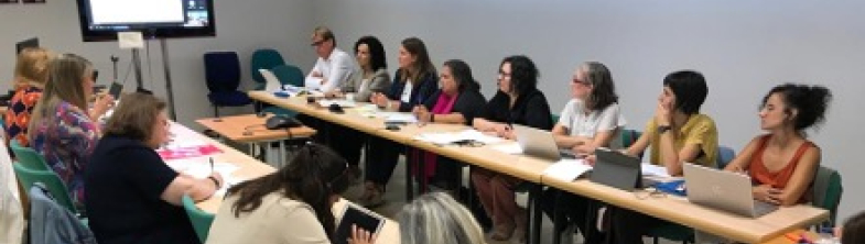 A group of people sit around a conference table, watching a presentation on a screen
