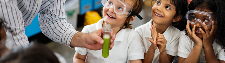 A teacher shows a test tube full of green liquid to three children wearing eye protection and smiling