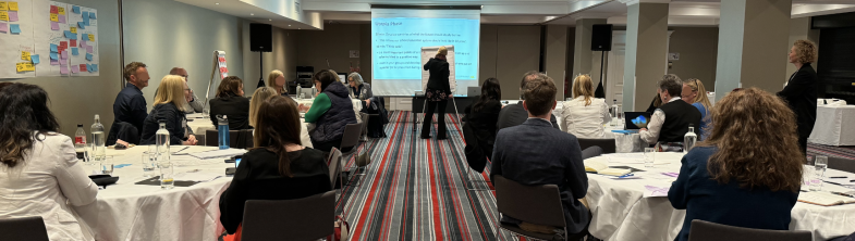 A person writes on a flip chart, in front of groups of people sitting around tables on either side