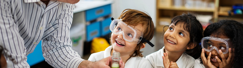 Children wearing safety goggles smile up at their teacher who is holding a test tube