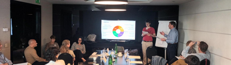 A young person stands in front of a screen making a presentation. A group of adults and young people sit listening around a conference table.