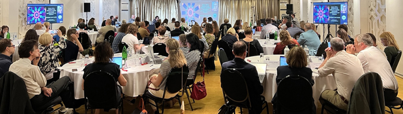 Participants at the bi-annual meeting sit around round tables and watch a presentation on a screen at the front of the room