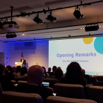 A screen which reads 'Opening remarks', with Pia Ahrenkilde Hansen standing at a lectern to the left