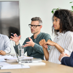 Three people sit at a conference table. The person in the middle is speaking