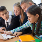 Three teenagers look at a laptop on a desk with their teacher