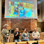 Four people sit in front of a presentation screen, one of them is speaking into a microphone