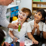 Children wearing safety goggles smile up at their teacher who is holding a test tube