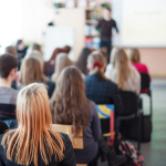 A group of people sit in rows, listening to someone speaking at the front of the room